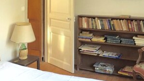 A bedroom with a bed, a bedside table with a lamp, a bookshelf filled with books, and a door.