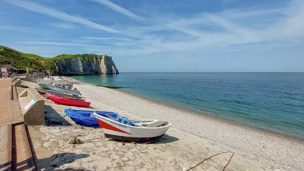 A beach with boats lined up on the shore, a cliff with greenery on top, and a clear blue sky.