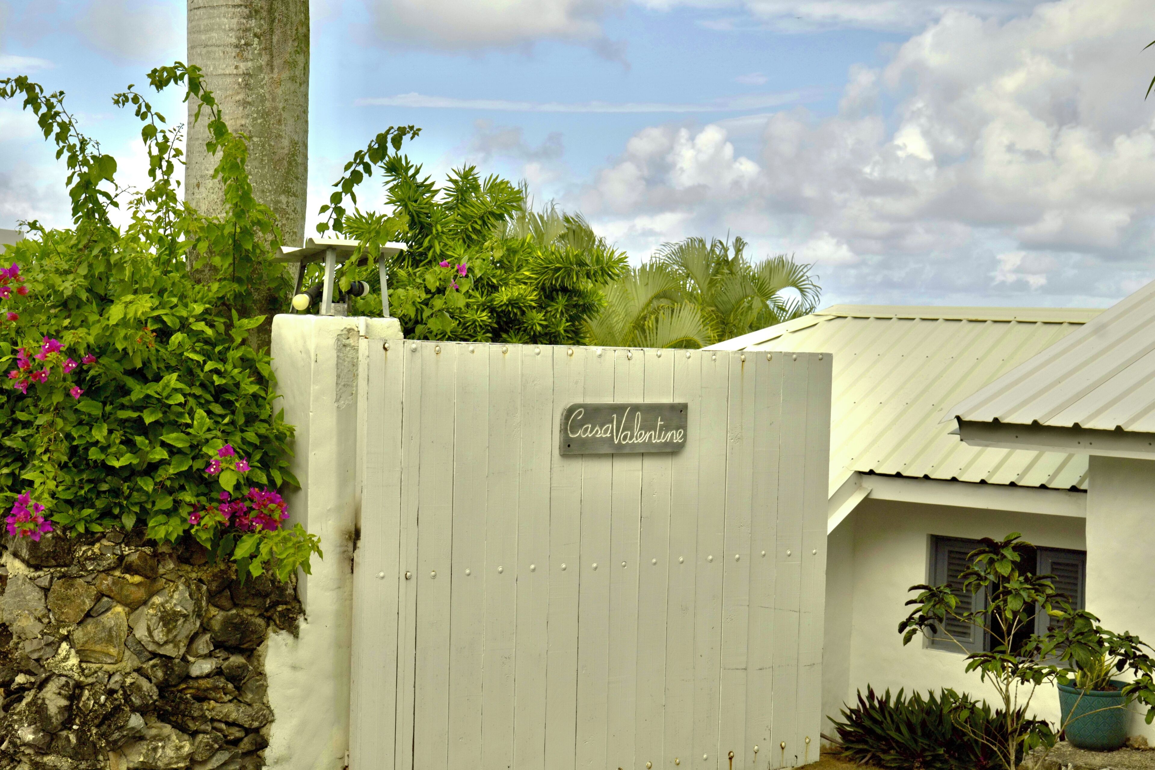 A white wooden gate with a sign reading "CasaValentine," a stone wall, and lush greenery including pink flowers and a palm tree.