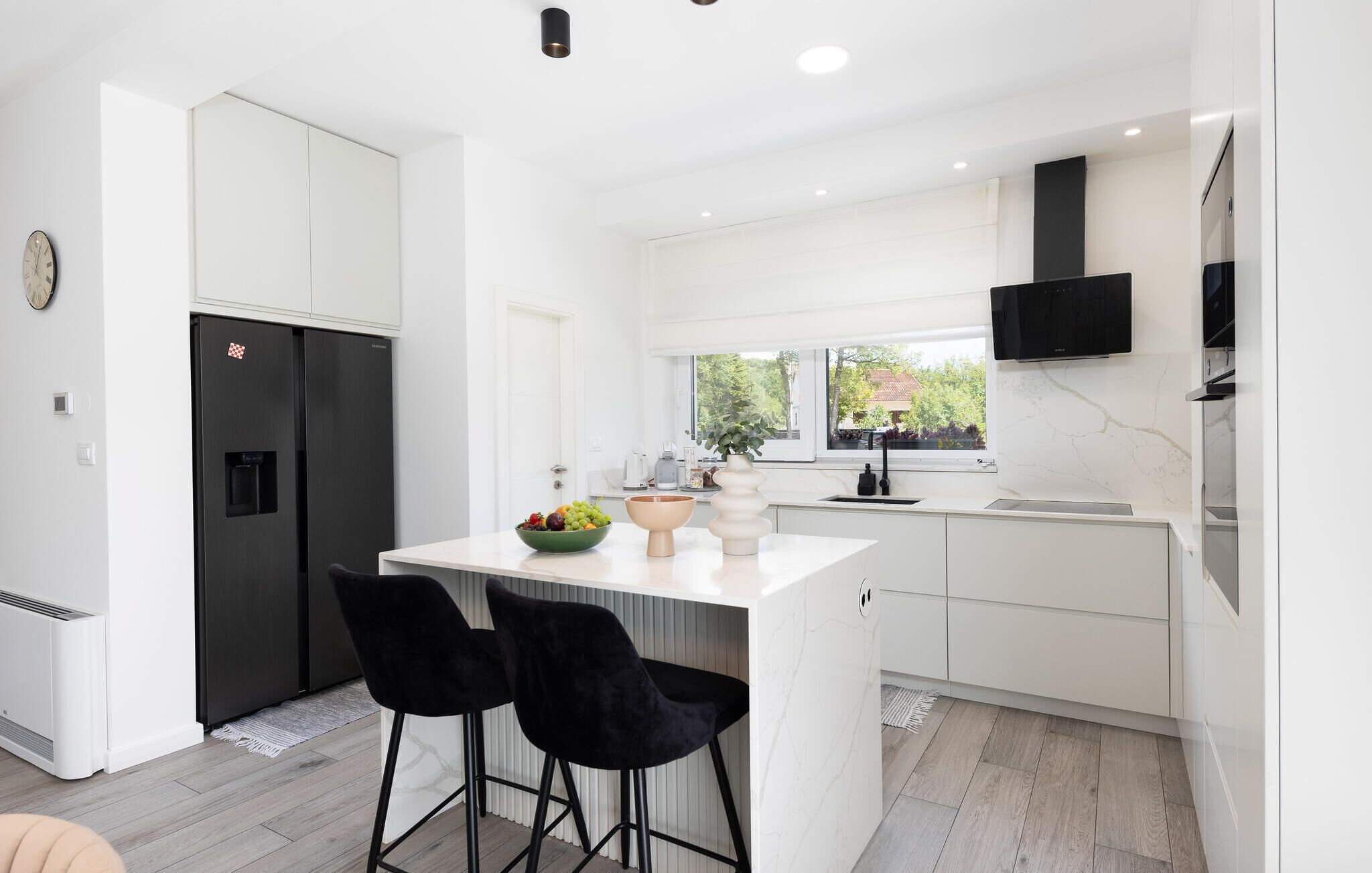 A modern kitchen with white cabinetry, a black refrigerator, a dining area with two black chairs, and a window with a view of greenery outside.