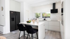 Une cuisine moderne avec des meubles blancs, un réfrigérateur noir, un coin repas avec deux chaises noires et une fenêtre donnant sur un paysage verdoyant.