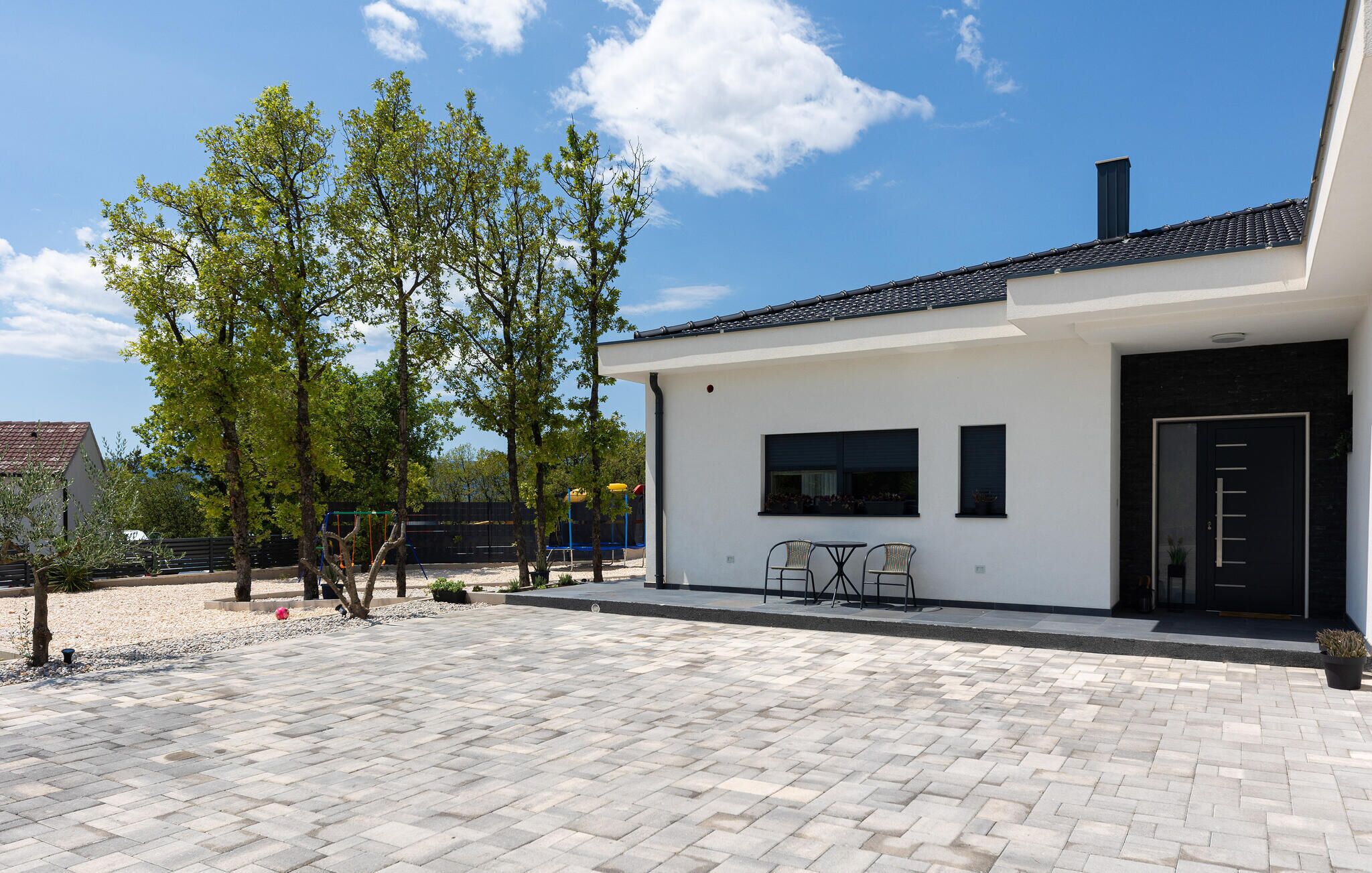 A modern house with a paved patio, a small outdoor seating area, and a playground in the background.