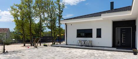 A modern house with a paved patio, a small outdoor seating area, and a playground in the background.