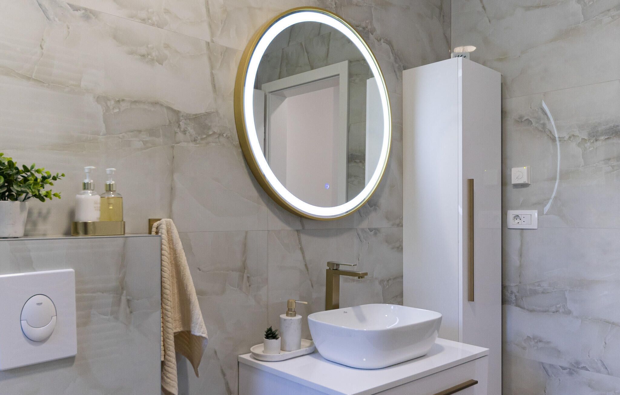A bathroom with a white vanity, a round mirror with a gold frame, a white sink, and a towel hanging on the wall.