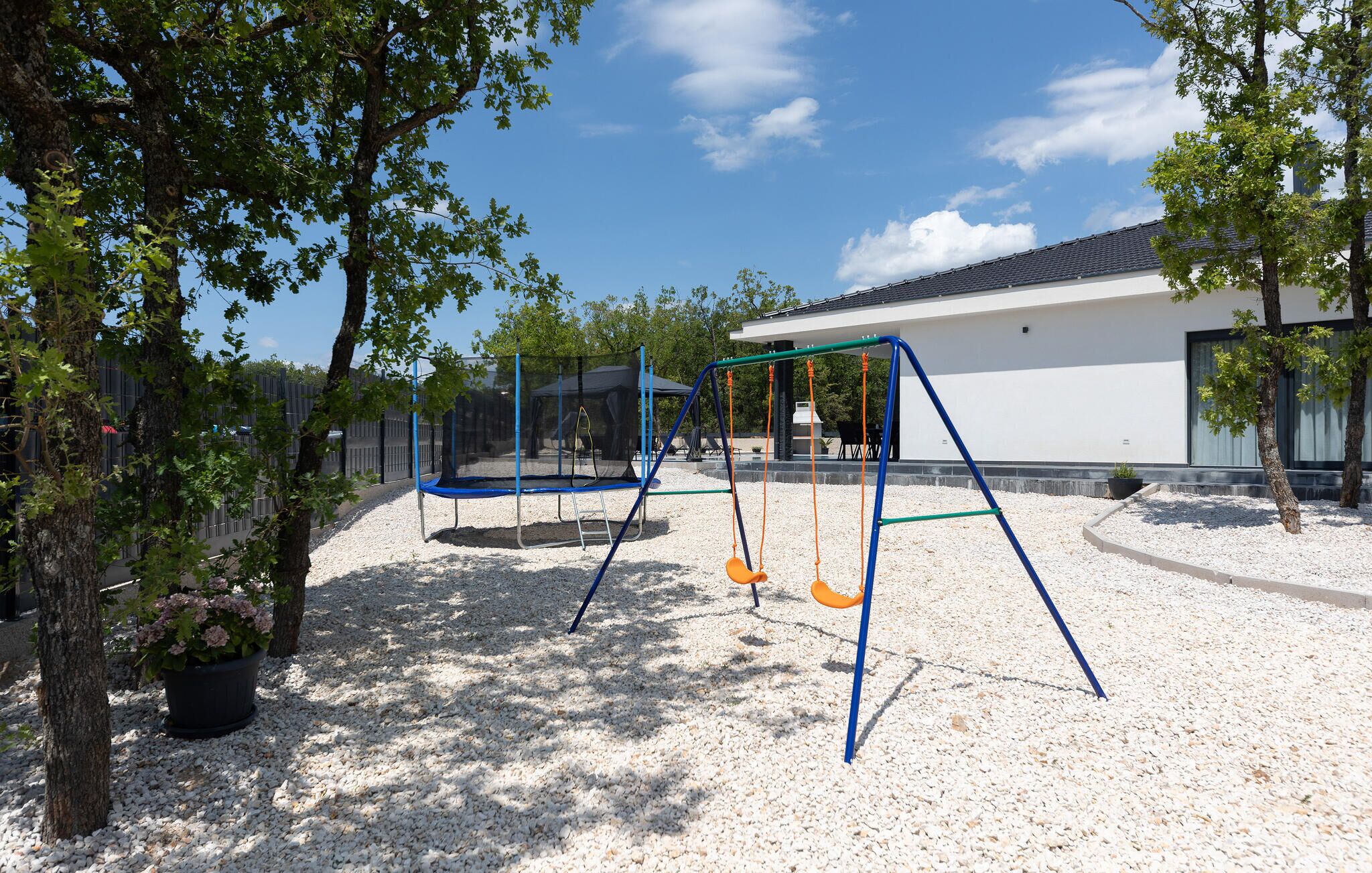 A playground with swings and a trampoline, surrounded by trees and a building in the background.