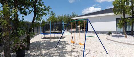 A playground with swings and a trampoline, surrounded by trees and a building in the background.