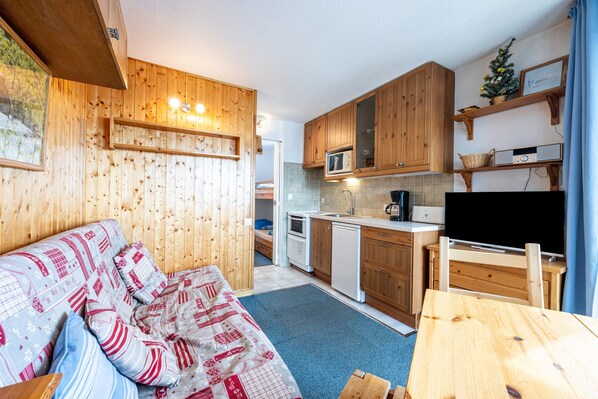 A cozy room with a bed, a wooden table, a kitchen with wooden cabinets, a microwave, and a television.
