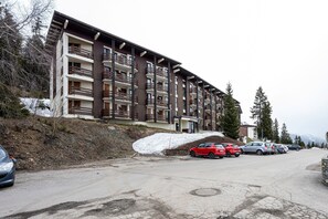 A multi-story building with balconies, a parking lot with several cars, and a snowy slope. - Studio "Les Bouquetins A" with Balcony and Valley View (South Facing) (La Rosière de Montvalezan)
