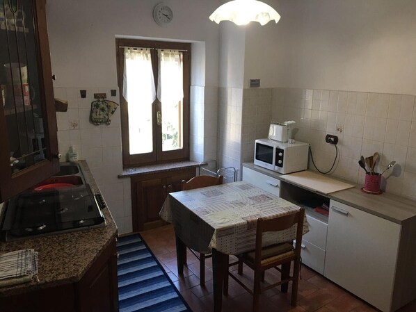 Une cuisine compacte équipée d’une cuisinière à gaz, d’un four à micro-ondes, d’une table à manger avec des chaises, d’une fenêtre avec des rideaux et d’une horloge au mur.