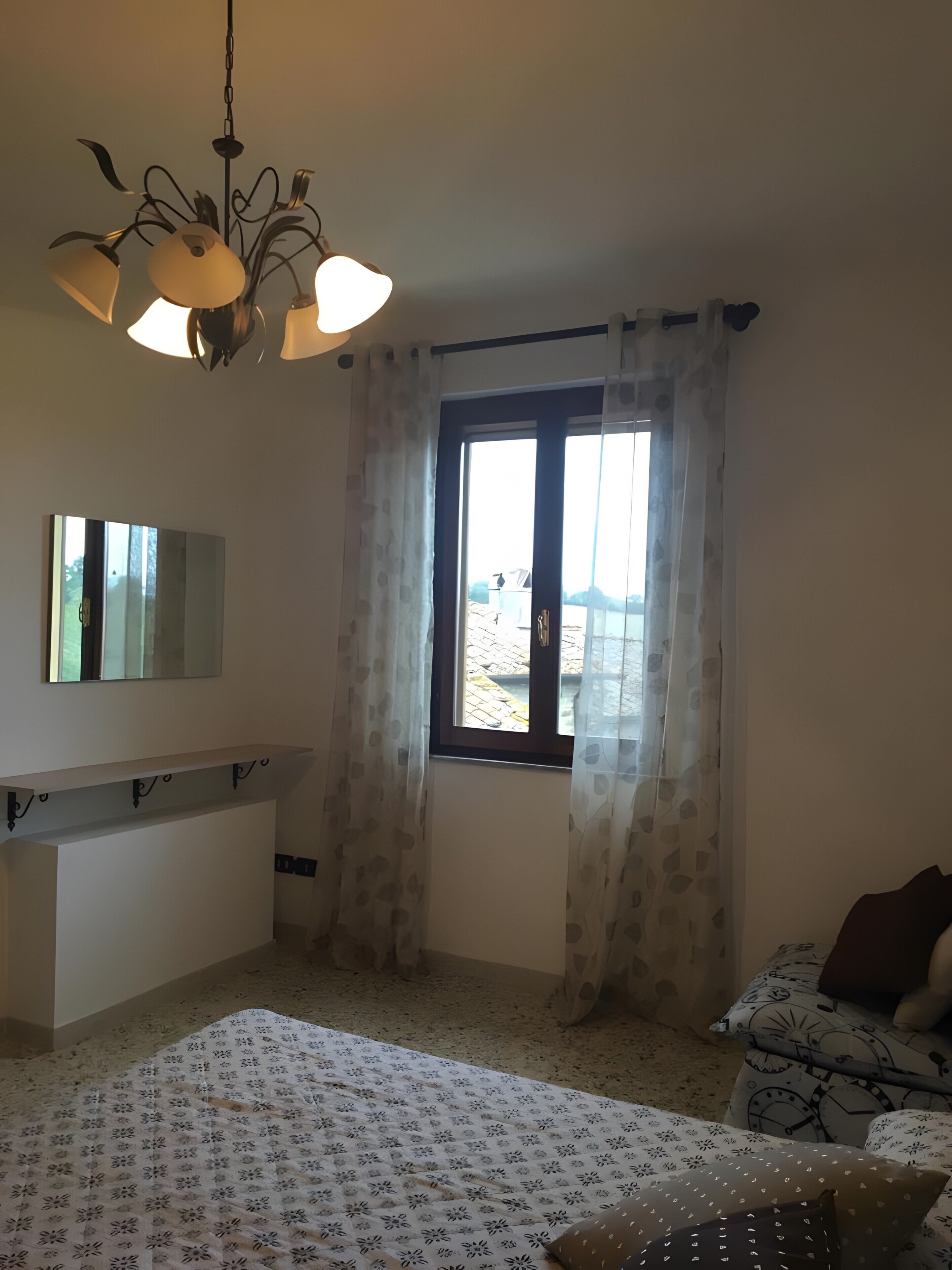 A bedroom with a bed, a bedside table, a mirror, a window with curtains, and a ceiling light fixture.