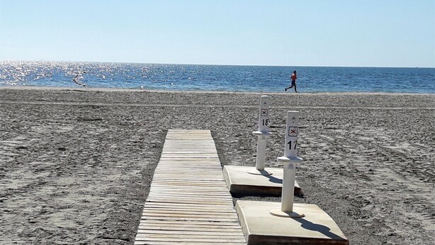 Ein hölzerner Weg führt zu einem Strand, wo zwei weiße Pfosten mit Nummern gekennzeichnet sind.