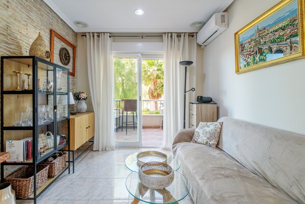 A living room with a sofa, a glass coffee table, a cabinet with decorative items, and a view through a glass door to an outdoor area.