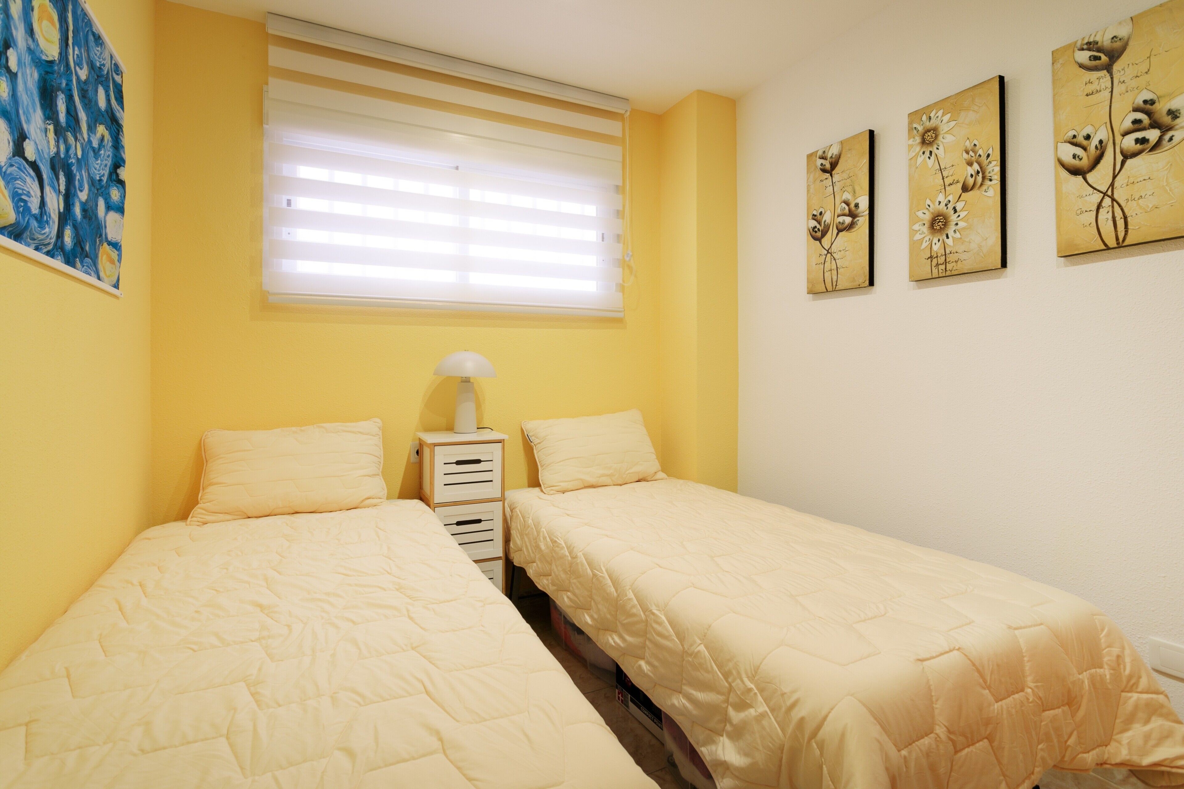 A room with two beds, a window with blinds, a bedside table with a lamp, and wall art.