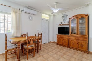 A dining area with a wooden table and chairs, a ceiling fan, a TV, and a cabinet with glass doors. - Bungalow 'Adelina En Santa Pola' with Private Terrace, Wi-Fi and Air Conditioning (Santa Pola)