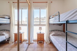 Quarto com beliches, mesa de cabeceira de madeira com abajur, e vista para edifícios através de grandes janelas.