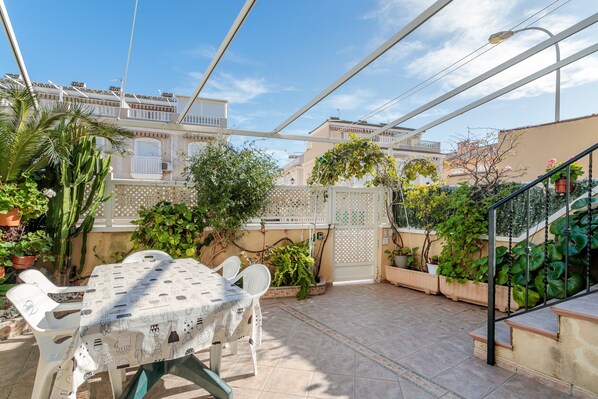 A covered patio with a table and chairs, surrounded by potted plants and a staircase.