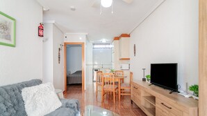 Uma sala de estar com sofá, mesa de centro, TV em um suporte de madeira, área de jantar com mesa e cadeiras, e uma cozinha ao fundo.