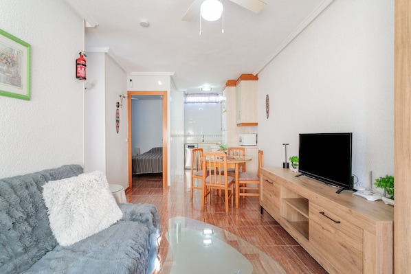 Un salon avec un canapé, une table basse, un téléviseur posé sur un meuble en bois, un coin repas avec une table et des chaises, et une cuisine à l’arrière-plan.