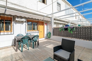 Une terrasse avec une table et des chaises, un fauteuil en osier, une porte en bois et une fenêtre avec des barreaux.