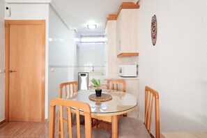 A dining area with a round glass table, wooden chairs, a potted plant, and a kitchen in the background.
