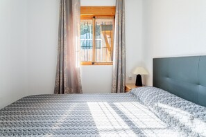 A bedroom with a bed, a window with curtains, a bedside table with a lamp, and a headboard.