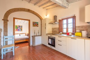 Une cuisine avec des meubles blancs, un fourneau, une fenêtre et une porte donnant sur une chambre à coucher.