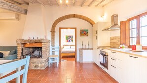 A kitchen with a stone fireplace, a dining area with a blue chair, and a view through an archway to a bedroom. - Apartment 'Casamoira' with Private Terrace, Wi-Fi and Air Conditioning (Trequanda)