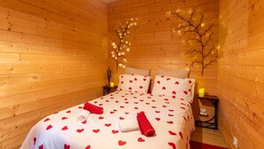 A cozy wooden room with a bed featuring heart-patterned bedding, a small bedside table with a lamp, and a decorative branch with lights.