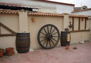 A courtyard with a large wooden wheel, two barrels, and potted plants against a wall with terracotta tiles and a window with wooden shutters.