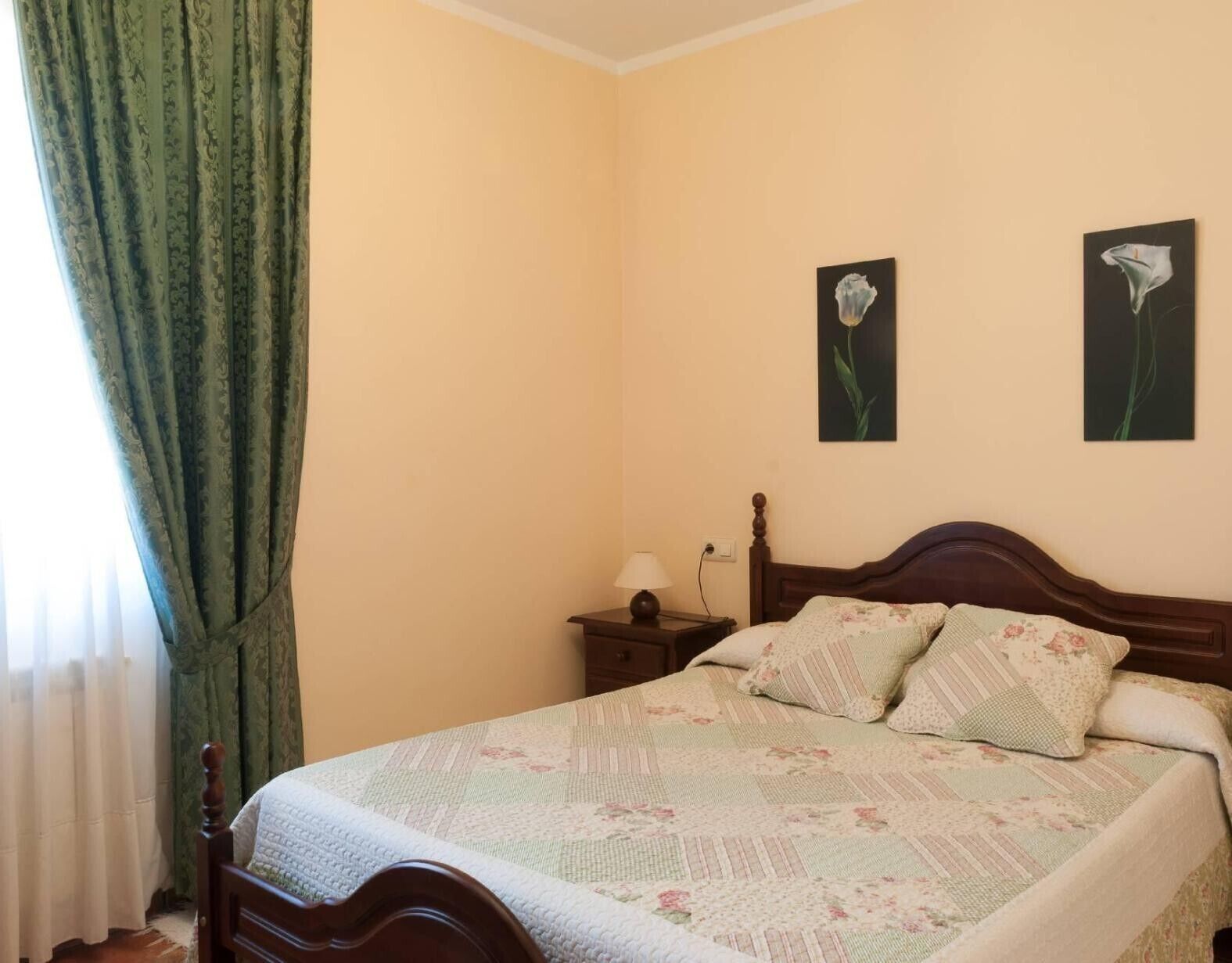 Un dormitorio con cama, mesita de noche, dos cuadros en la pared y una ventana con cortinas.