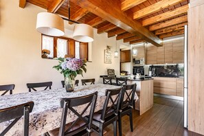 A dining area with a table set for six, wooden chairs, a vase with flowers, and a kitchen in the background. - Country House 'Hortensia' with Mountain View, Private Terrace and Wi-Fi (Asturias)