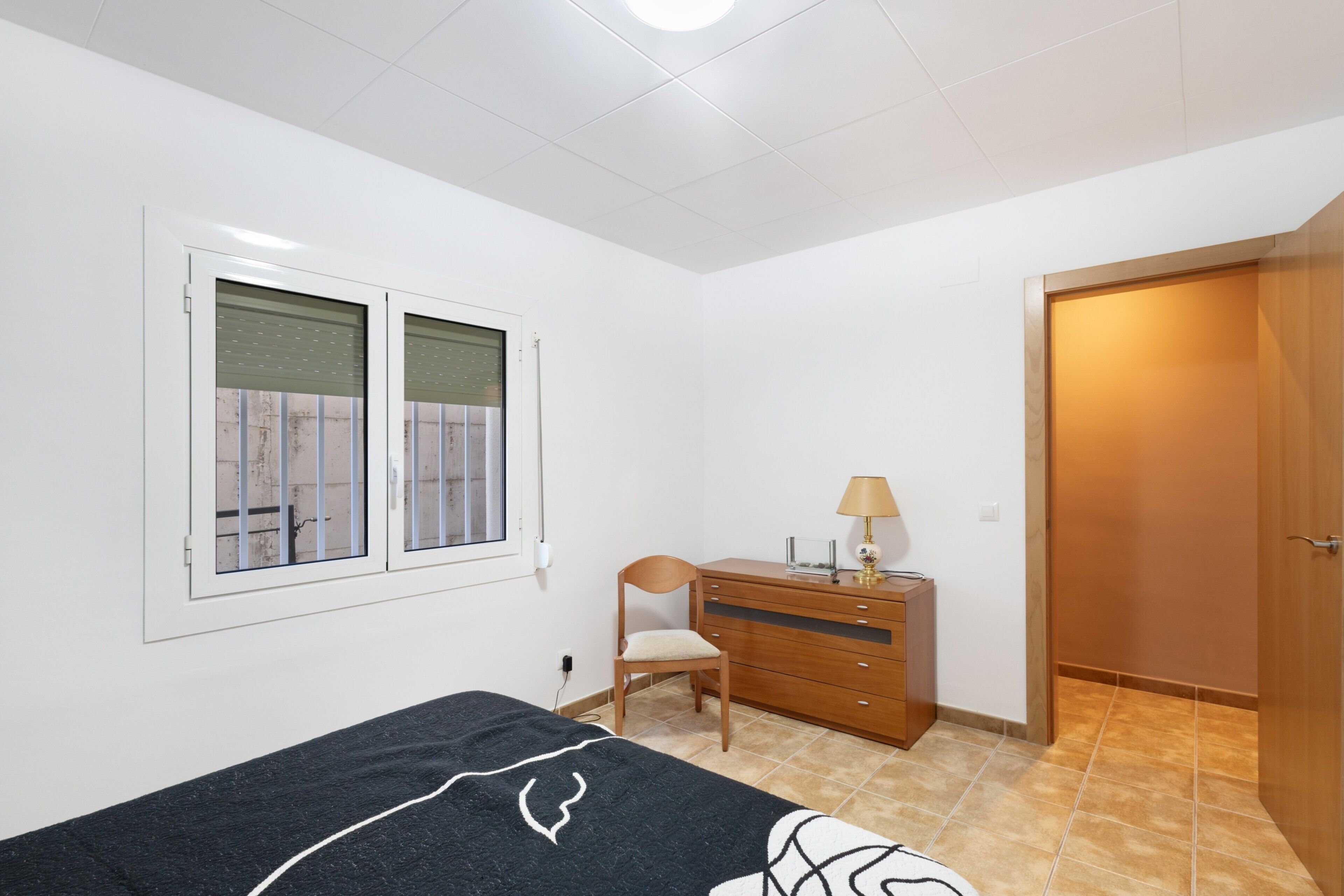 A bedroom with a bed, a wooden dresser, a chair, a window with blinds, and a door.