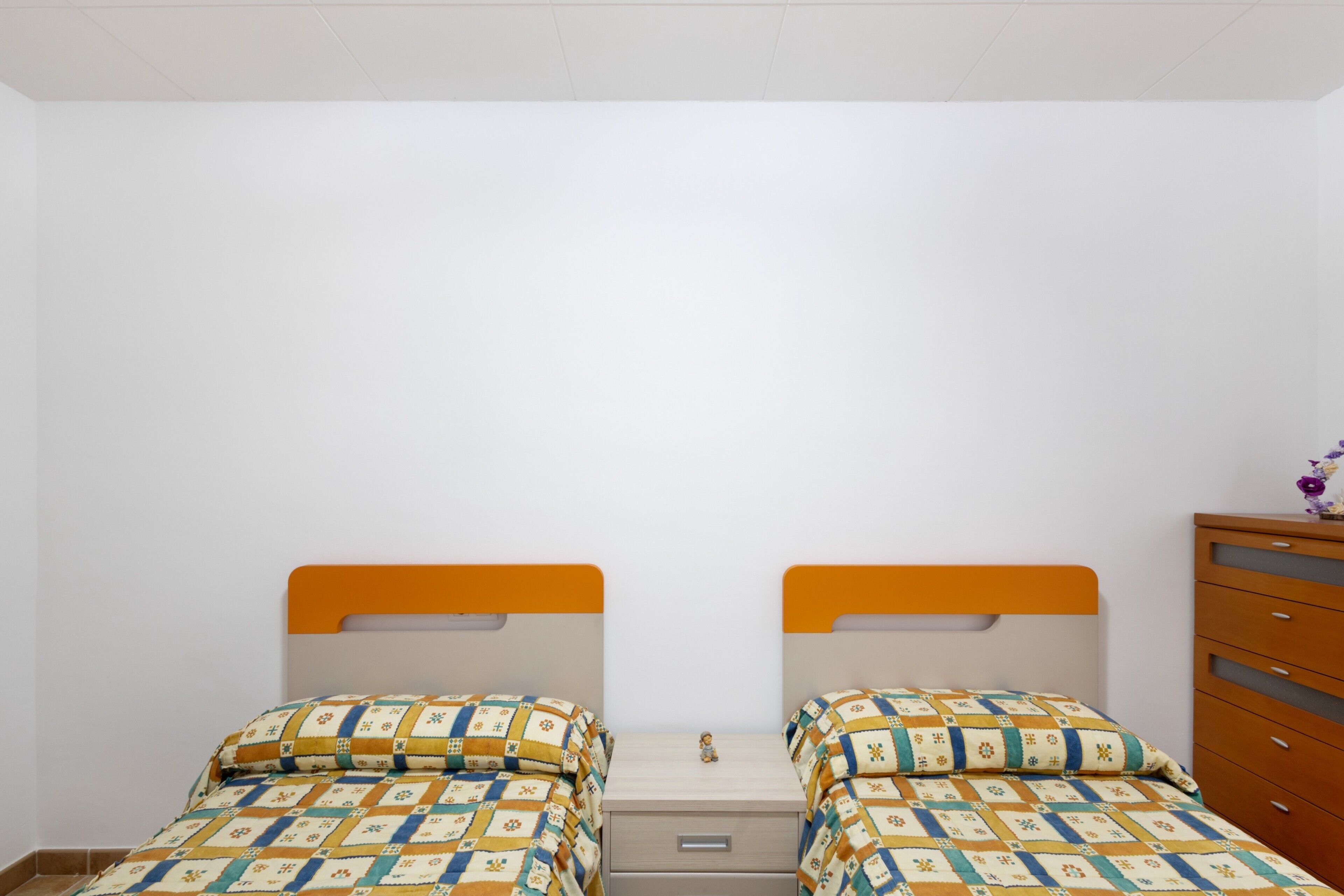A room with two beds, each with a patterned bedspread and an orange headboard. There is a wooden dresser with drawers and a small vase with flowers on top.