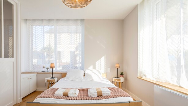Ein Schlafzimmer mit einem Bett, Nachttischen, einem Schreibtisch und einem Fenster mit leichtem Vorhang.