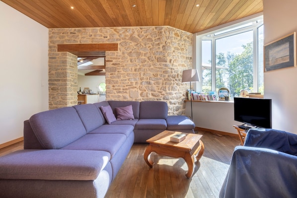 Un salon moderne avec un mur en pierre, un plafond en bois, un canapé d’angle, une table basse, un téléviseur et une bibliothèque.