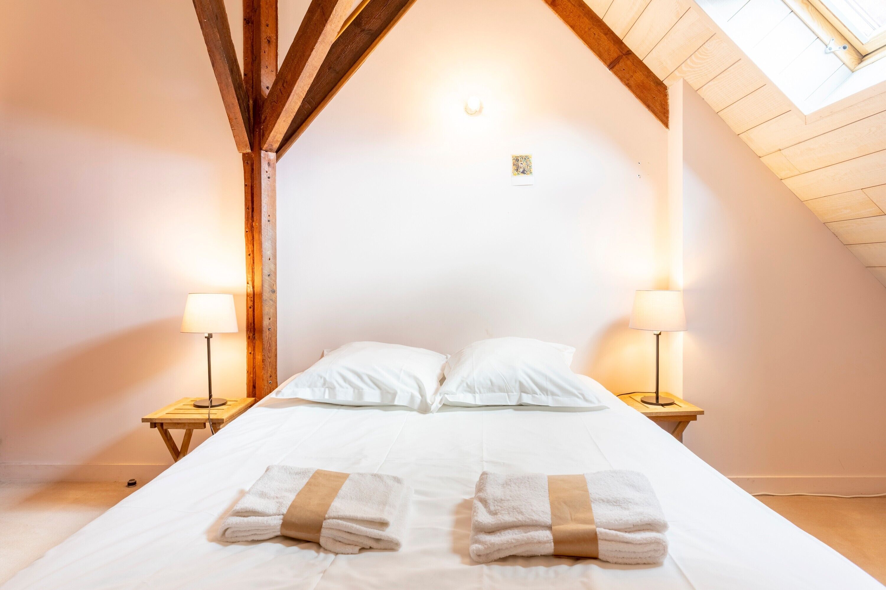 Une chambre avec un plafond en pente, deux lampes de chevet, un lit avec du linge de lit blanc et une lucarne.