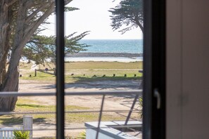 Blick durch ein Fenster auf einen Strand mit Sandfläche, einem Holzzaun und einem großen Baum.