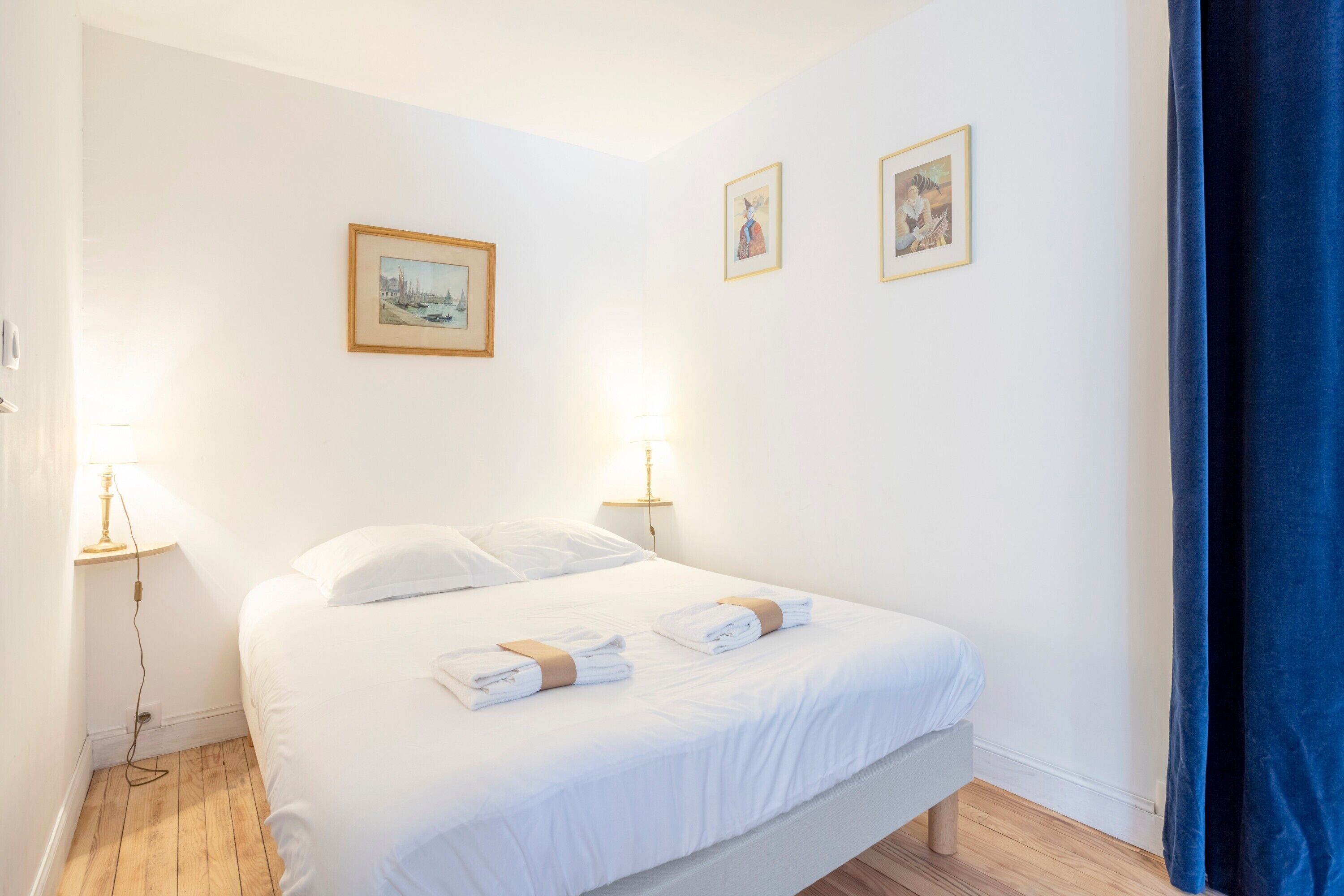 Hotellihuone, jossa on sänky, kaksi yöpöytävalaisinta, yöpöytä ja kolme kehystettyä kuvaa seinällä.