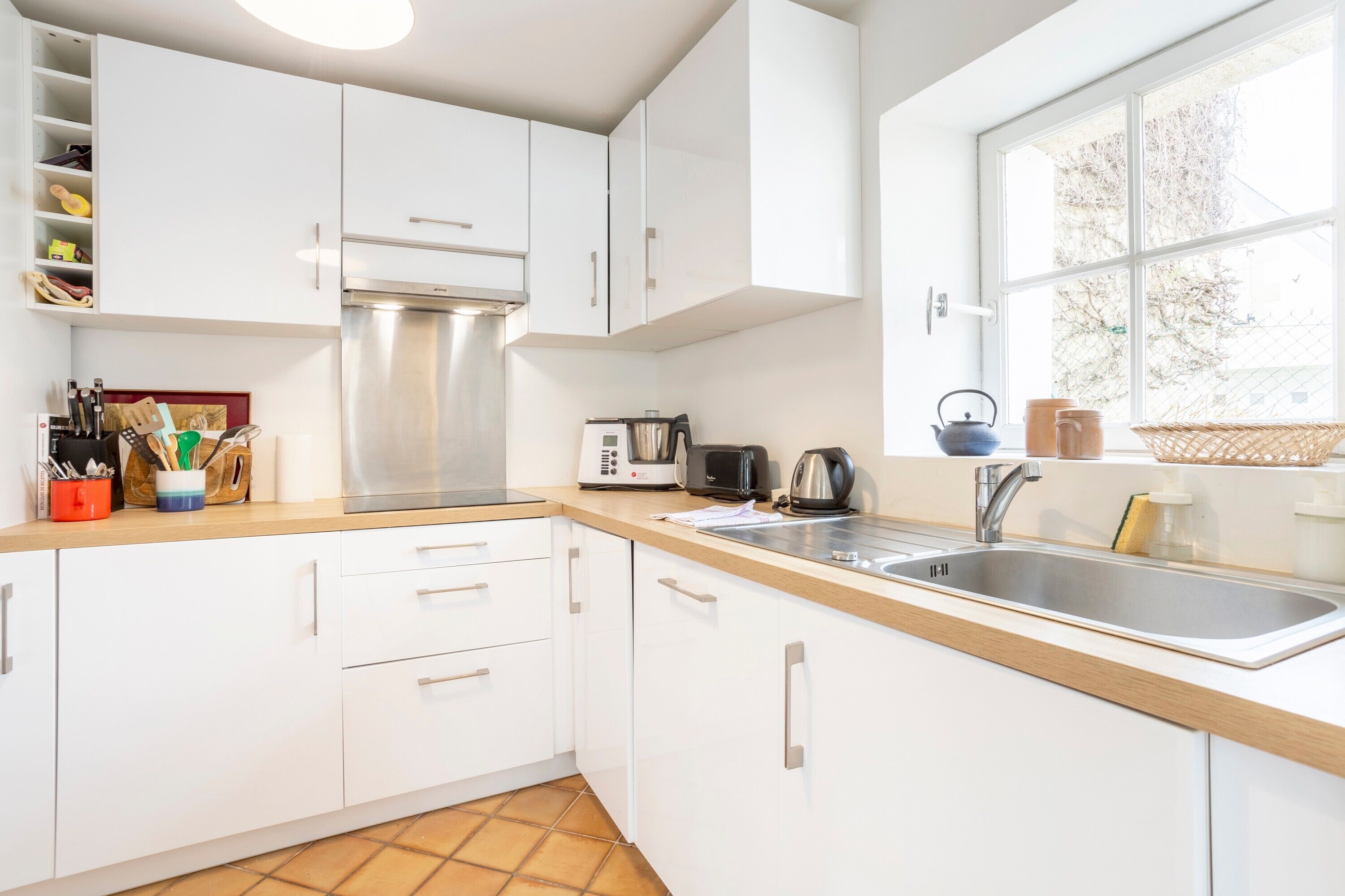 Une cuisine moderne avec des meubles blancs, une hotte de cuisinière en acier inoxydable, un évier et une fenêtre donnant sur des arbres.