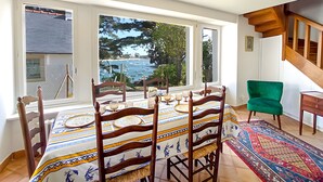A dining area with a table set for four, large windows with a view of the sea, a green armchair, and a wooden staircase. - L'Estuaire – River View, 20 Meters from Shops (Combrit)