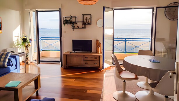 Een woonkamer met een televisie, bank en een eethoek, met uitzicht op een balkon en de zee.
