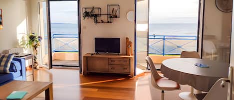 Sala de estar con televisor, sofá y comedor, con vista a un balcón al mar.