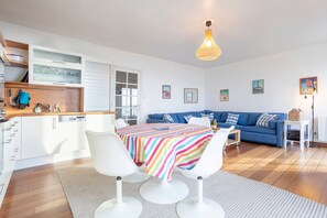 Une cuisine moderne avec des meubles blancs, une table à manger recouverte d’une nappe à rayures colorées et un canapé bleu dans l’espace salon.