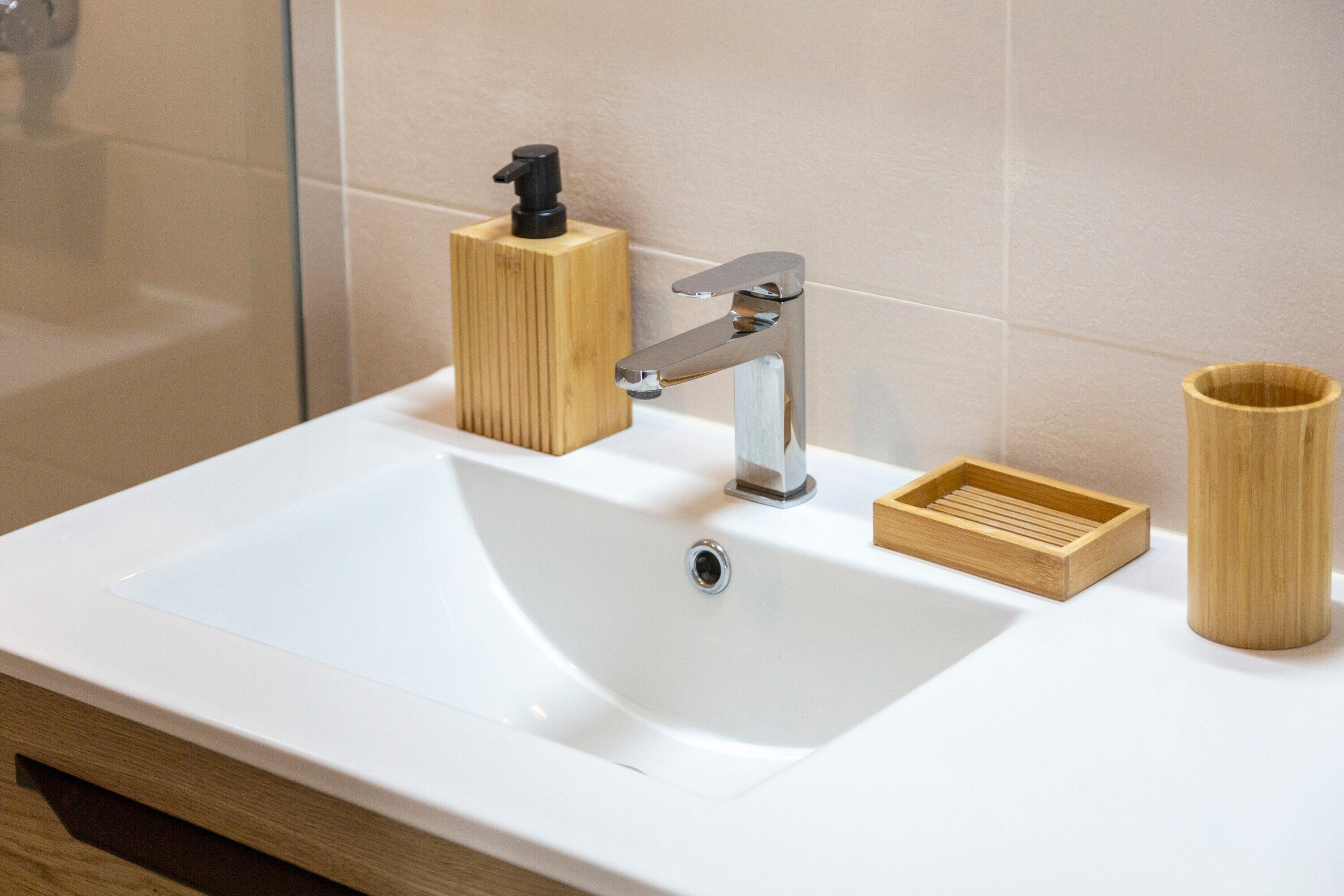 Une salle de bain avec un lavabo blanc, un distributeur de savon en bois, un gobelet en bambou et un plateau en bois.