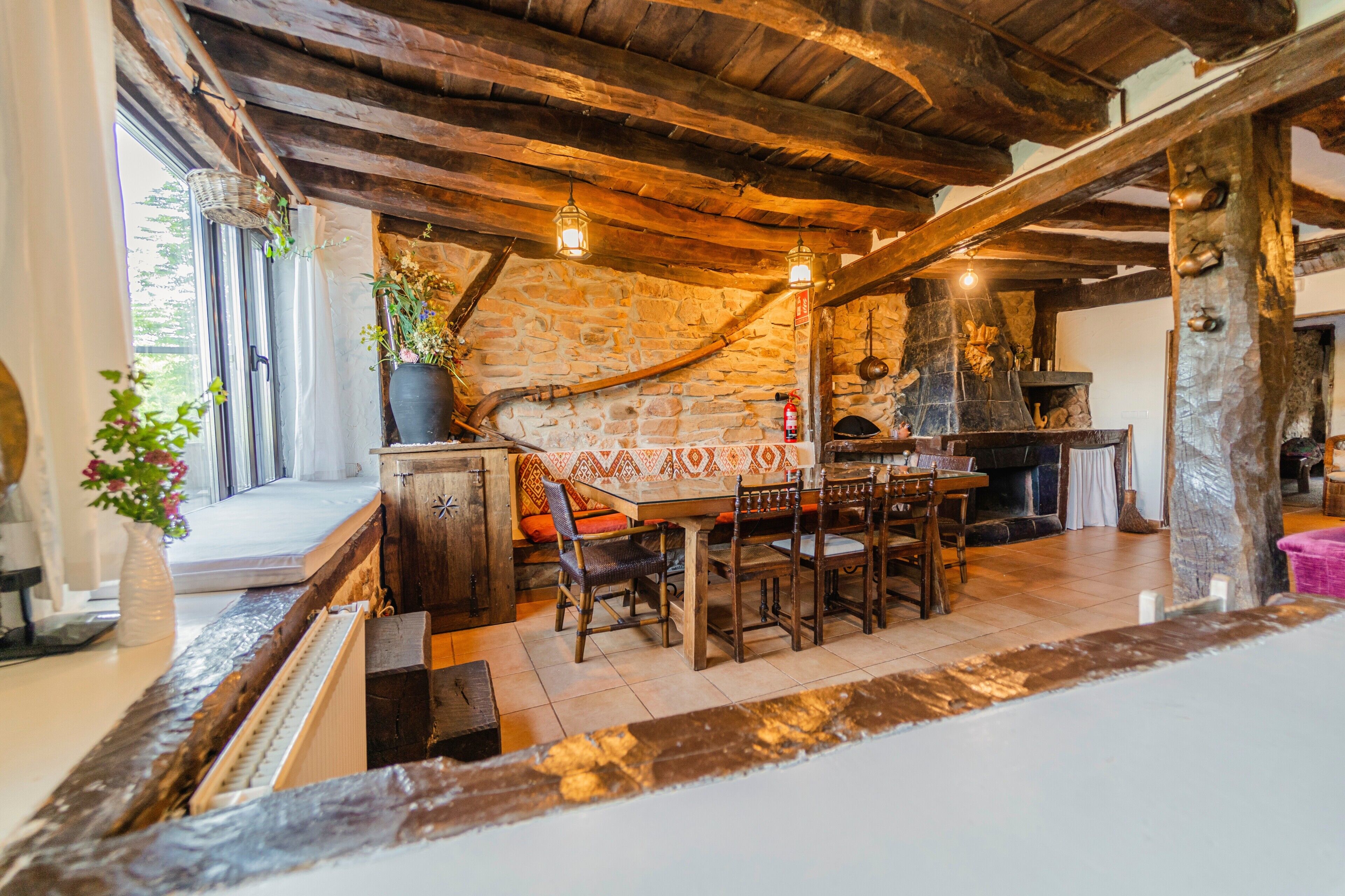 A rustic interior with exposed wooden beams, a stone wall, a fireplace, and a dining area with wooden furniture.