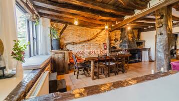 A rustic interior with exposed wooden beams, a stone wall, a fireplace, and a dining area with wooden furniture.