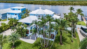 A large, modern house with multiple blue and white sections, surrounded by lush greenery and palm trees, near a body of water.