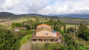 Une maison en pierre avec un toit de tuiles, entourée d’une végétation luxuriante et avec des collines en arrière-plan.