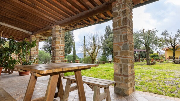 Une table et un banc en bois, placés sous une pergola en pierre et en bois, offrent une vue sur une zone herbeuse et des arbres.