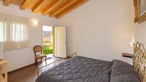 A bedroom with a bed, a chair, a wooden ceiling, a door leading outside, and a wall-mounted light fixture.